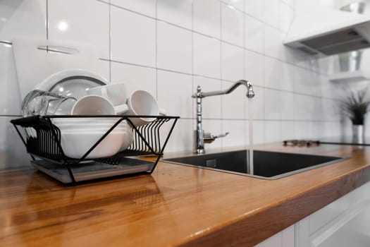 Budget and lightweight antimicrobial dish drainer with drain board at modern scandinavian kitchen. Dish rack holds many dishes and cups against wooden countertop, white wall tiles, sink and faucet
