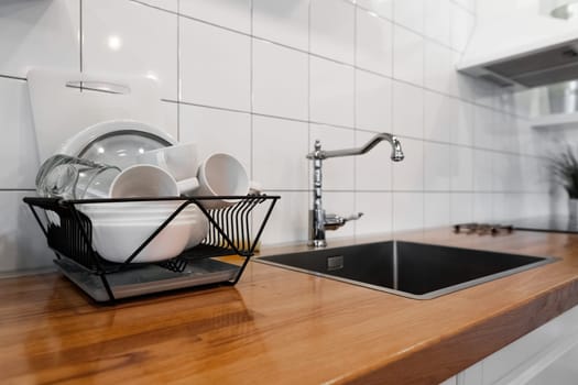 Budget and lightweight antimicrobial dish drainer with drain board at modern scandinavian kitchen. Dish rack holds many dishes and cups against wooden countertop, white wall tiles, sink and faucet