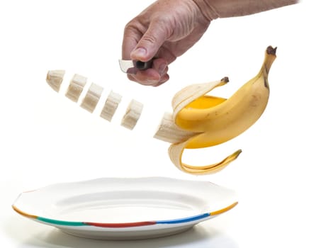A human hand holding a knife and slicing a half peeled banana with the slices suspended midair over a plate