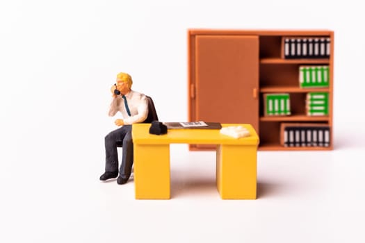 Miniature people businessman holding a telephone sitting behind his work desk. Macro shot of Tiny people business concept.