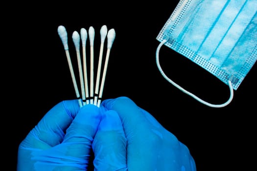 A close up of hands wearing gloves to a medical personal holding throat swabs next to a medical face mask on a black background.