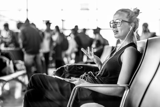 Casual sporty young blond female traveler talking on her cell phone while waiting to board a plane at the departure gates at the asian airport terminal.