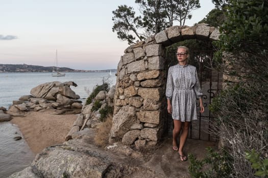 Beautiful woman in luxury summer dress enjoying peaceful seascape of Porto Rafael bay at Mediterranean sea of Costa Smeralda, Sardinia, Italy at dusk.