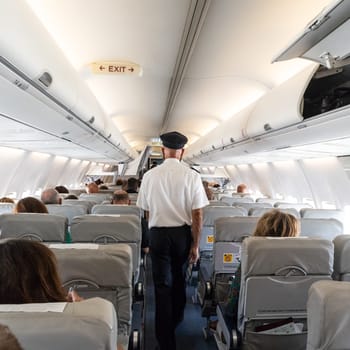 Interior of commercial airplane with flight attandant serving passengers on seats during flight. Steward in uniform walking the aisle.square composition.