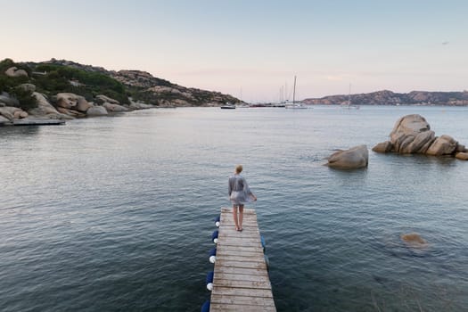Beautiful woman in luxury summer dress standing on wooden pier enjoying peaceful seascape at dusk. Female traveler stands on a wooden pier in Porto Rafael, Costa Smeralda, Sardinia, Italy.