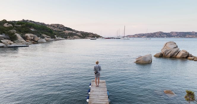 Beautiful woman in luxury summer dress standing on wooden pier enjoying peaceful seascape at dusk. Female traveler stands on a wooden pier in Porto Rafael, Costa Smeralda, Sardinia, Italy.