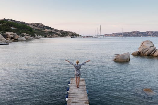Beautiful woman standing on wooden pier arms rised enjoying peaceful seascape at dusk. Female traveler stands on a wooden pier in Porto Rafael, Costa Smeralda, Sardinia, Italy.