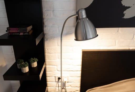 Steel downbridge floor lamp with a long arm and opaque shade near bed in black and white modern bedroom. Metal Task Lamp with an adjustable reading light. Stylish bedside lamp in scandinavian interior