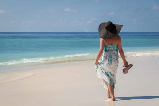 Maldives, young woman walking along the beach with sunhat and high heels on the hand.