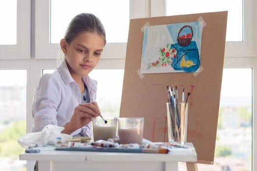 girl sits behind an easel and washes her brush in a glass of water