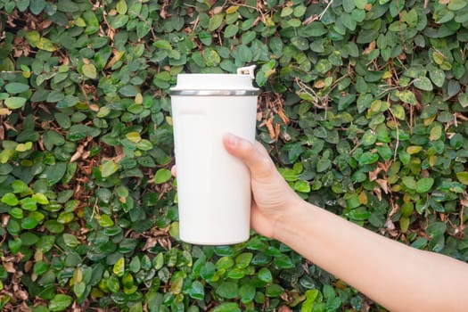 Aluminum thermos mug on hand with green leaves wall, stock photo