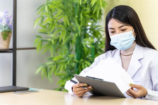 Portrait of asian female doctor wear protective face mask sit in her office room in hospital clinic and reading on patience file before checking patience. Health care Concept.