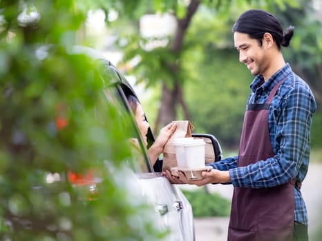 Waiter giving hot coffee cup with disposable tray and bakery bag through car window to customer at drive thru service station. Drive thru is popular service after coronavirus covid-19 pandemic.