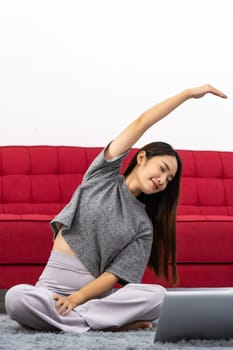 Asian woman using laptop for online coaching to do exercising while sitting in lotus position . Active young woman doing stretching exercise in living room at home. Fit firm good mental health concept