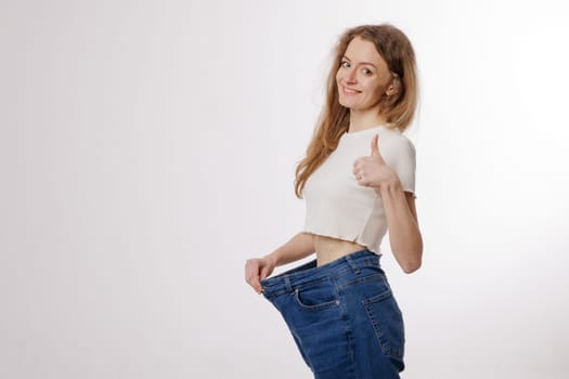 Happy woman showing weight loss success by wearing oversized jeans, smiling, and giving a thumbs-up on a white background.