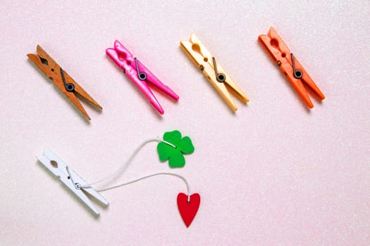 Colorful set of five wooden and plastic clothespins on a light pink glitter sparkle background with space. A red heart and a green lucky clover leaf are attached to the white wooden clip. Topview.