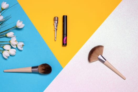Professional makeup brushes, lipsticks and spring flowers flat lay on a bright pink, yellow and blue background with copy space. Template for cosmetics product display montage or advertising text. Beauty and skincare concept. Top view.