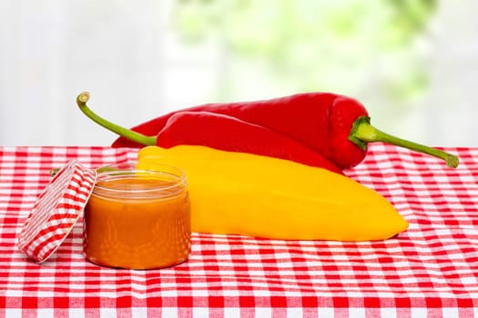 Closeup of a jar with home made classical sauce of fresh paprika with tomatoes, horseradish, garlic and basil and three paprika on a red checkered tablecloth over bright background. Template for food and product display montage. Macro.