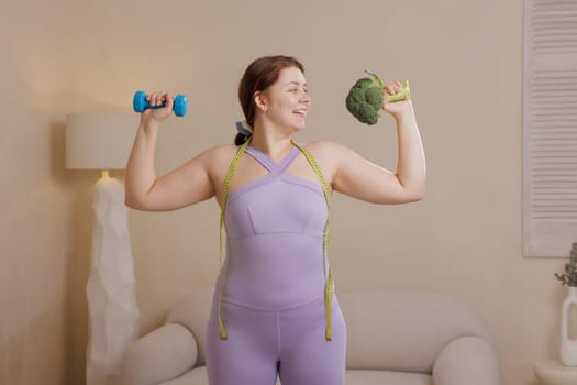 Happy overweight woman in sportswear combining fitness and healthy eating holding dumbbell and broccoli symbolizing well-being, weight loss and body positivity.