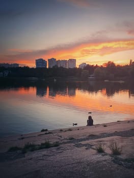 Moody and idyllic evening scene with a little girl silhouette, feeding the wild ducks, sitting near the lake in the park. Wonderful summer sunset with colorful reflection on the calm water surface.