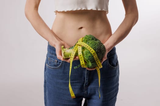 Slim woman holding broccoli wrapped in a measuring tape, symbolizing healthy eating, weight loss, diet, and nutrition on a white background.