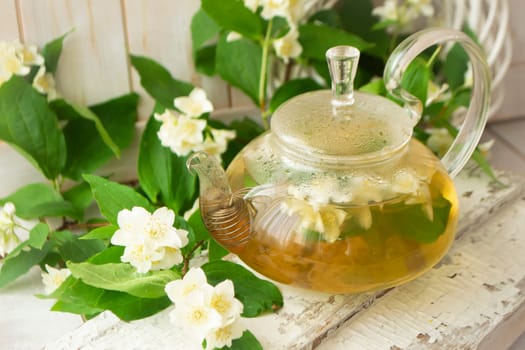 glass teapot with hot tea and jasmine flowers. branches of blooming jasmine on shabby background.