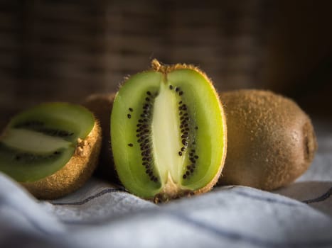 Close-up of a group of kiwis, one of them cut in half. Dark background with a wicker basket. On a white tablecloth with blue lines.