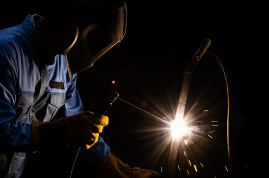 Shielded metal arc welding with sparks. A man welder in blue uniform with welding helmet and yellow leather gloves. Industrial working with personnel protective equipment concept.