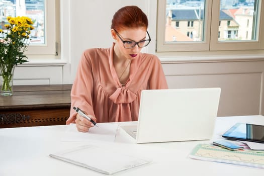 Young businesswoman reads statistics at the desk on the computer with red hair and glasses in the office