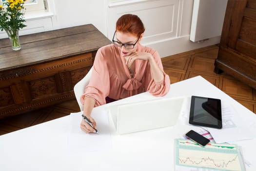 Young businesswoman reads statistics at the desk on the computer with red hair and glasses in the office