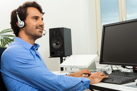 Attractive young man with headset in the office at customer service operator phone service cheerful with blue shirt