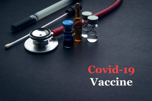 COVID-19 or CORONAVIRUS VACCINE text with stethoscope, medical swab and vial on black background. Covid-19 or Coronavirus concept.