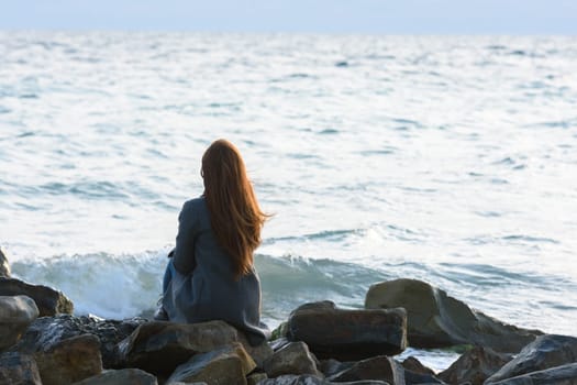 A lonely girl sits on the rocks by the sea and looks into the distance