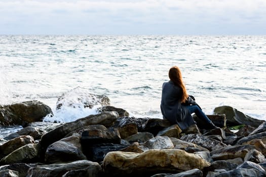 A girl sits on stony stones by the sea and looks into the distance