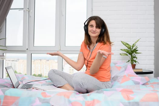 The girl spread her hands in surprise, sitting in headphones and with a laptop on the bed at home