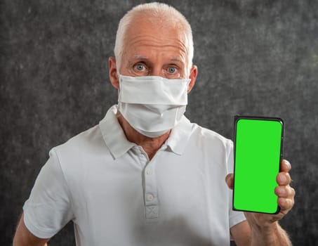 a senior man in a medical mask showing smartphone with green screen. Concept of the danger of coronavirus