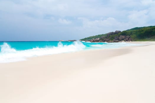 White sand beach with some waves and blue sky in the far a hill with green plants