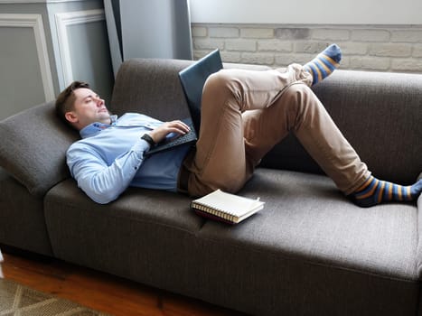 A man lies at home on the couch and works on a laptop. Remote work concept.