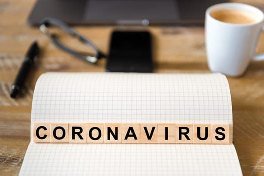 Closeup on notebook over wood table background, focus on wooden blocks with letters making Coronavirus writing. Business concept with laptop, glasses, pen and mobile phone in a defocused background