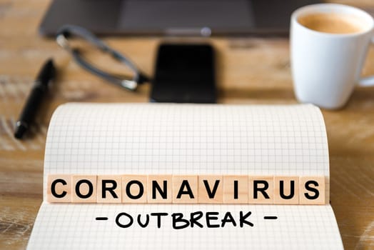 Closeup on notebook over wood table background, focus on wooden blocks with letters making Coronavirus Outbreak writing. Business concept with laptop and mobile phone in a defocused background