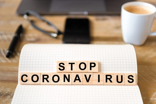 Closeup on notebook over wood table background, focus on wooden blocks with letters making Stop Coronavirus writing. Business concept with laptop and mobile phone in a defocused background