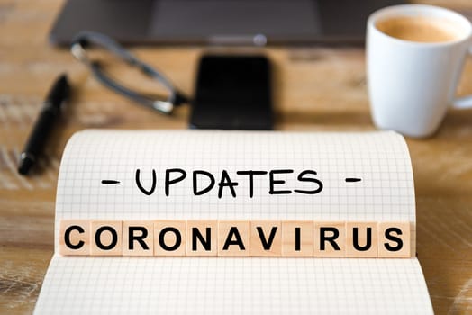 Closeup on notebook over wood table background, focus on wooden blocks with letters making Coronavirus Covid-19 updates writing. Business concept with laptop and mobile phone in a defocused background