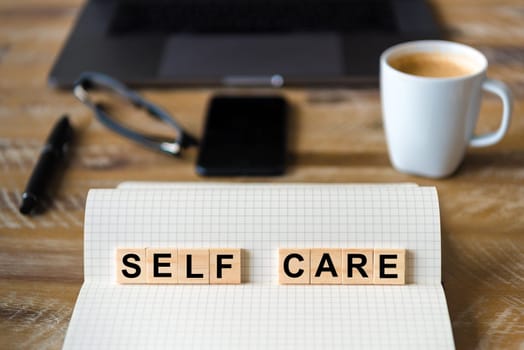 Closeup on notebook over wood table background, focus on wooden blocks with letters making Self Care writing. Business concept with laptop and mobile phone in a defocused background