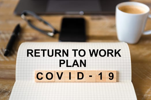 Closeup on notebook over wood table background, focus on wooden blocks with letters making Covid-19 Return To Work Plan writing. Business concept with laptop and mobile phone in defocused background