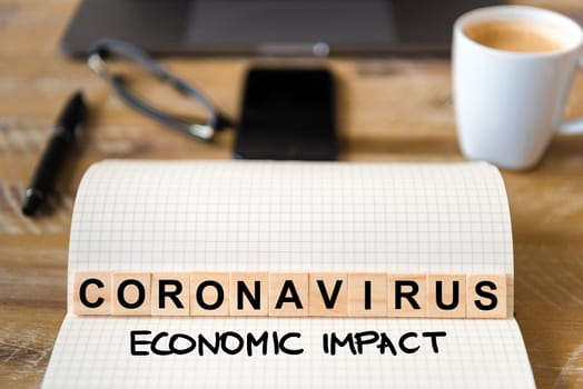 Closeup on notebook over wood table background, focus on wooden blocks with letters making Coronavirus Economic Impact writing. Business concept with laptop and mobile phone in a defocused background