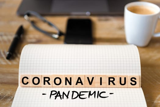 Closeup on notebook over wood table background, focus on wooden blocks with letters making Coronavirus Pandemic writing. Business concept with laptop and mobile phone in a defocused background