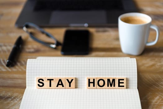Closeup on notebook over wood table background, focus on wooden blocks with letters making Stay Home writing. Business concept with laptop and mobile phone in a defocused background