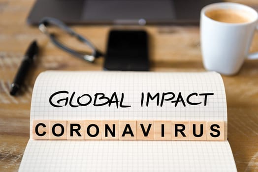 Closeup on notebook over wood table background, focus on wooden blocks with letters making Coronavirus Global Impact writing. Business concept with laptop and mobile phone in a defocused background