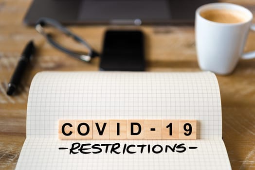 Closeup on notebook over wood table background, focus on wooden blocks with letters making Covid-19 restrictions writing. Business concept with laptop and mobile phone in a defocused background