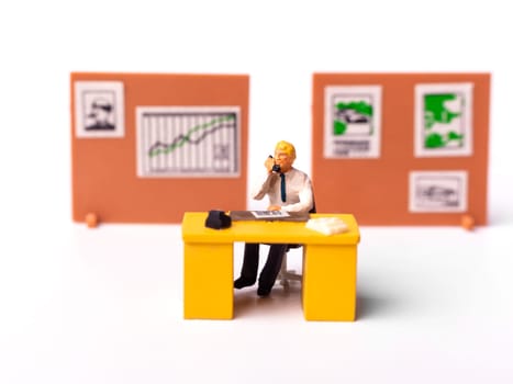Miniature people businessman holding telephone sitting behind his work desk with graph on the background. Macro shot of Tiny people business concept.